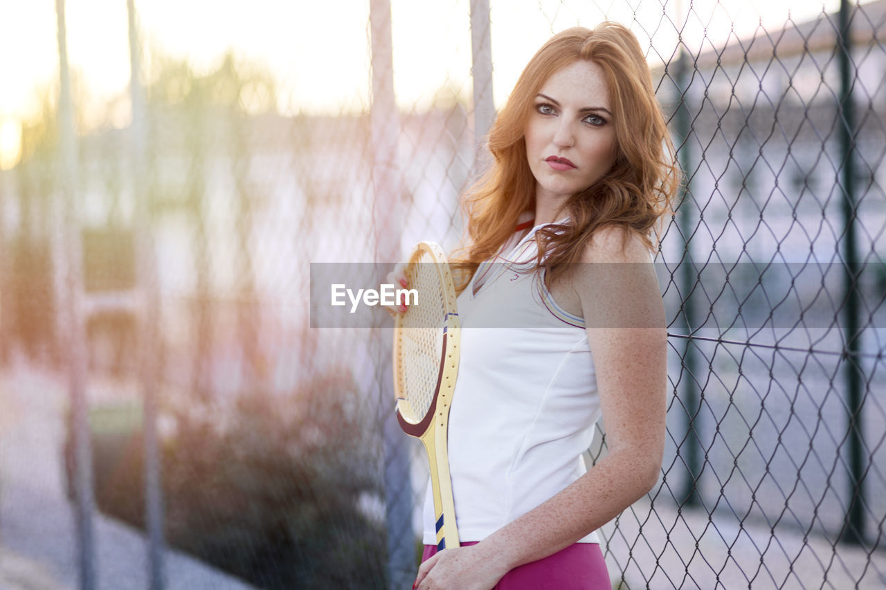 Portrait of woman holding badminton racket while standing against fence