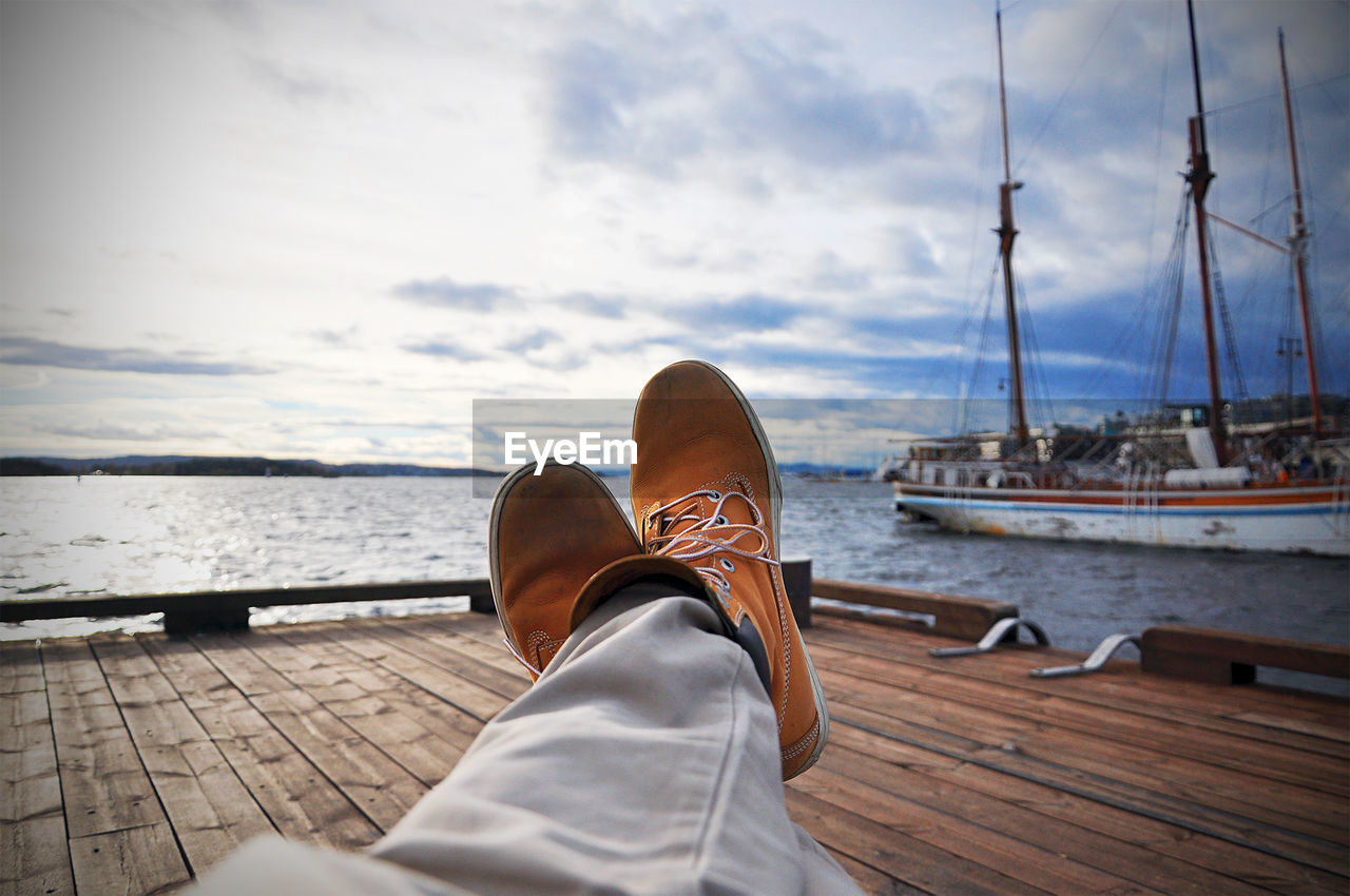 Low section of man resting on jetty by boats moored at lake against sky