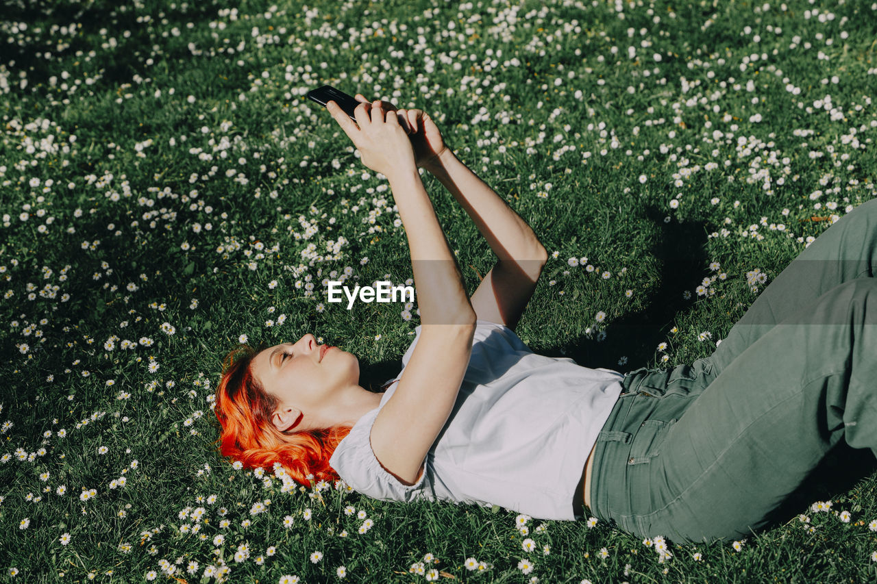 Side view of delighted redhead female lying on grass on lawn and messaging on mobile phone in summer
