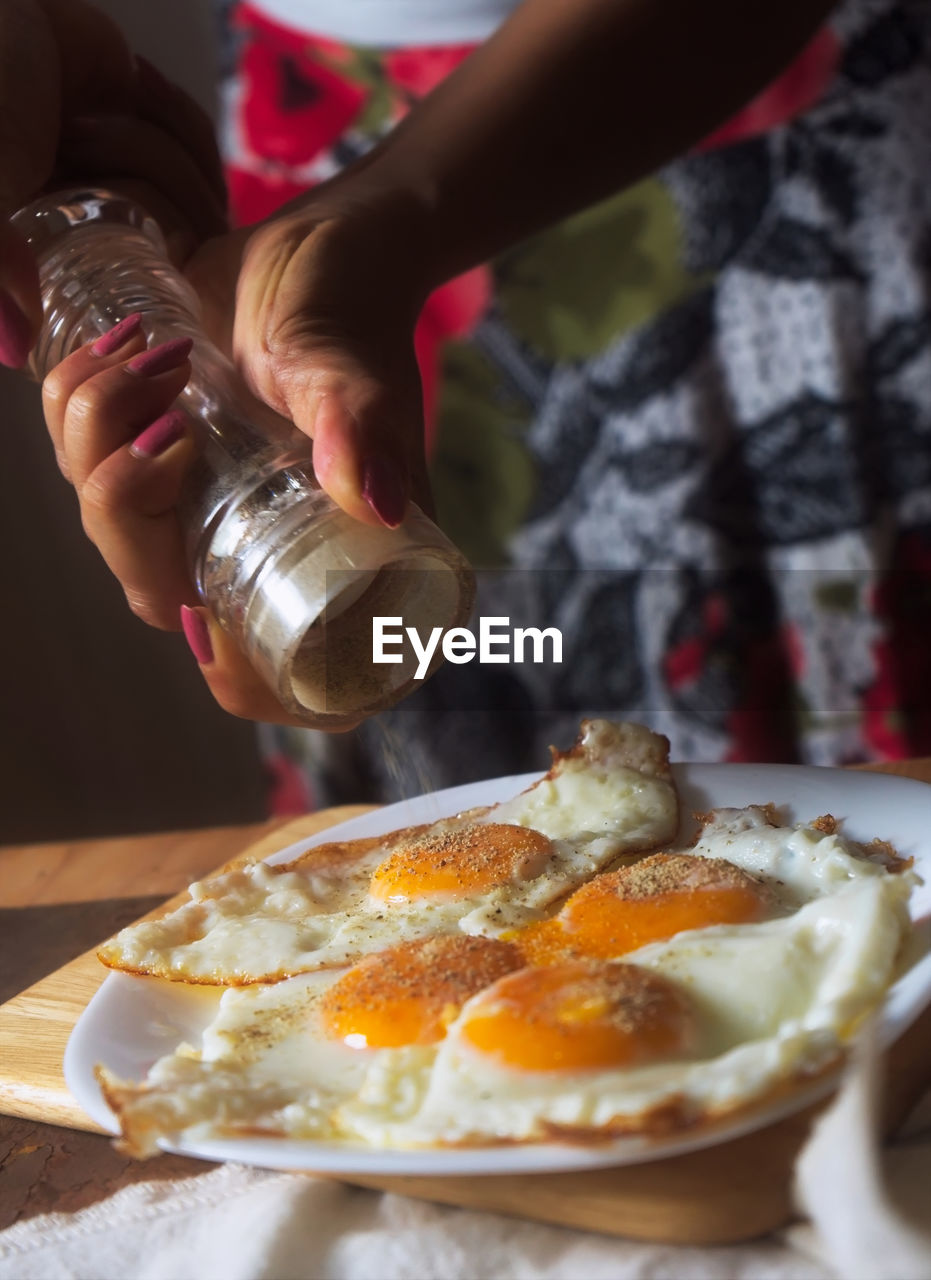 Cropped hands of woman sprinkling pepper on fried eggs at home