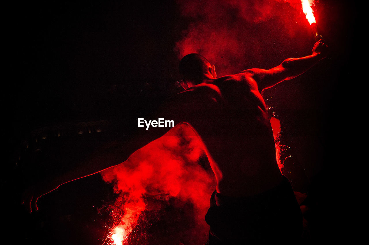 Rear view of shirtless male performer holding red distress flares