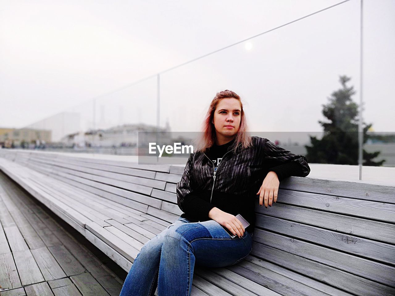 Young woman sitting on bench against sky