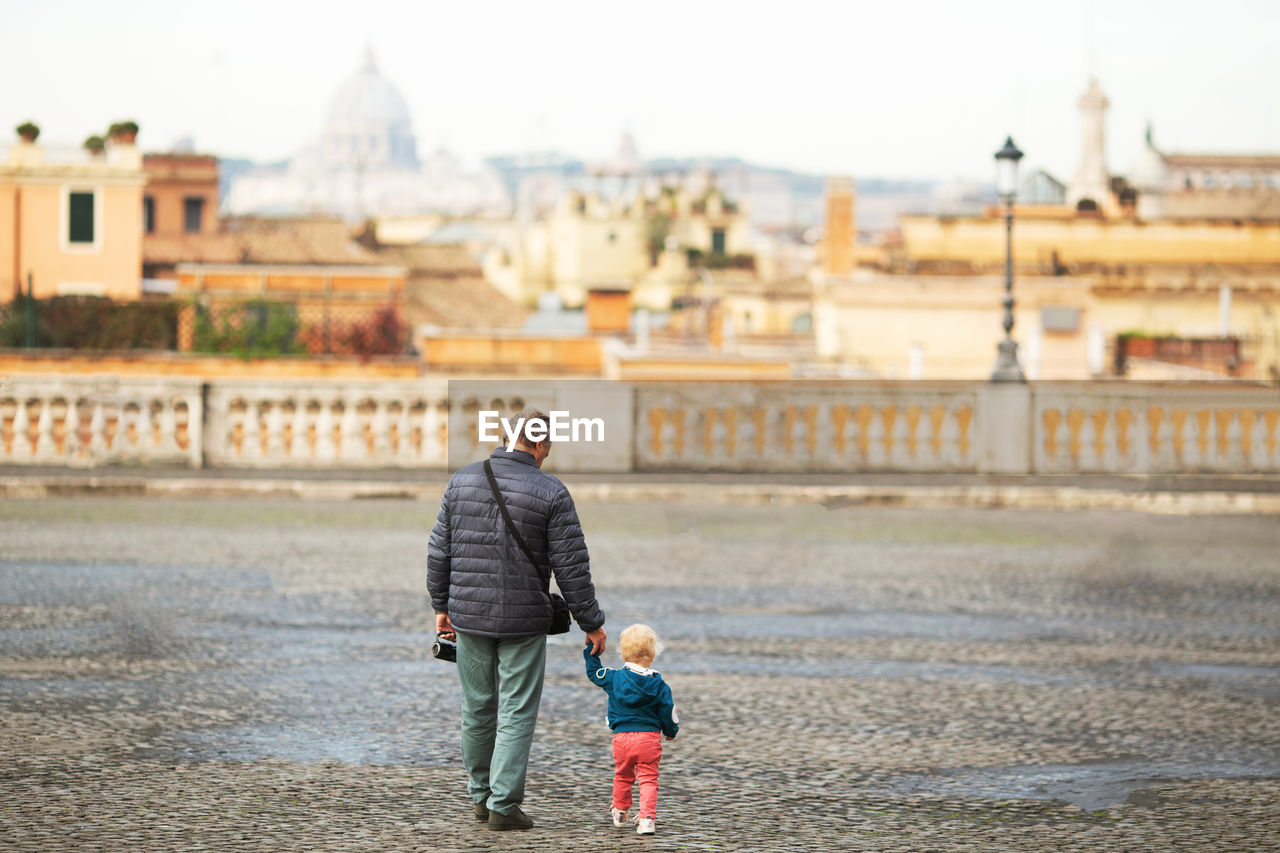 Rear view of grandfather holding granddaughter hand while walking on road