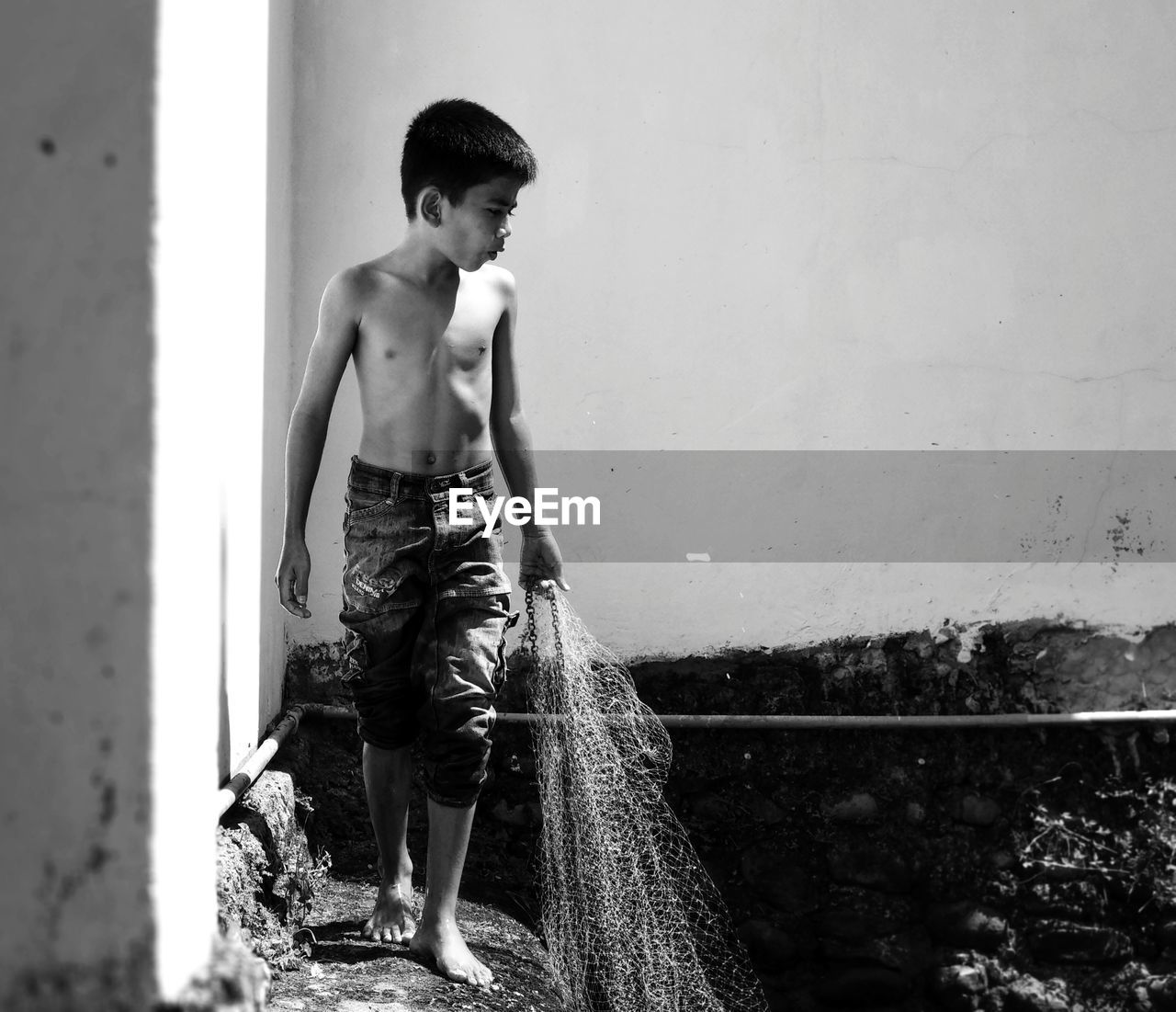 Full length of shirtless boy looking away while holding fishing net against sky