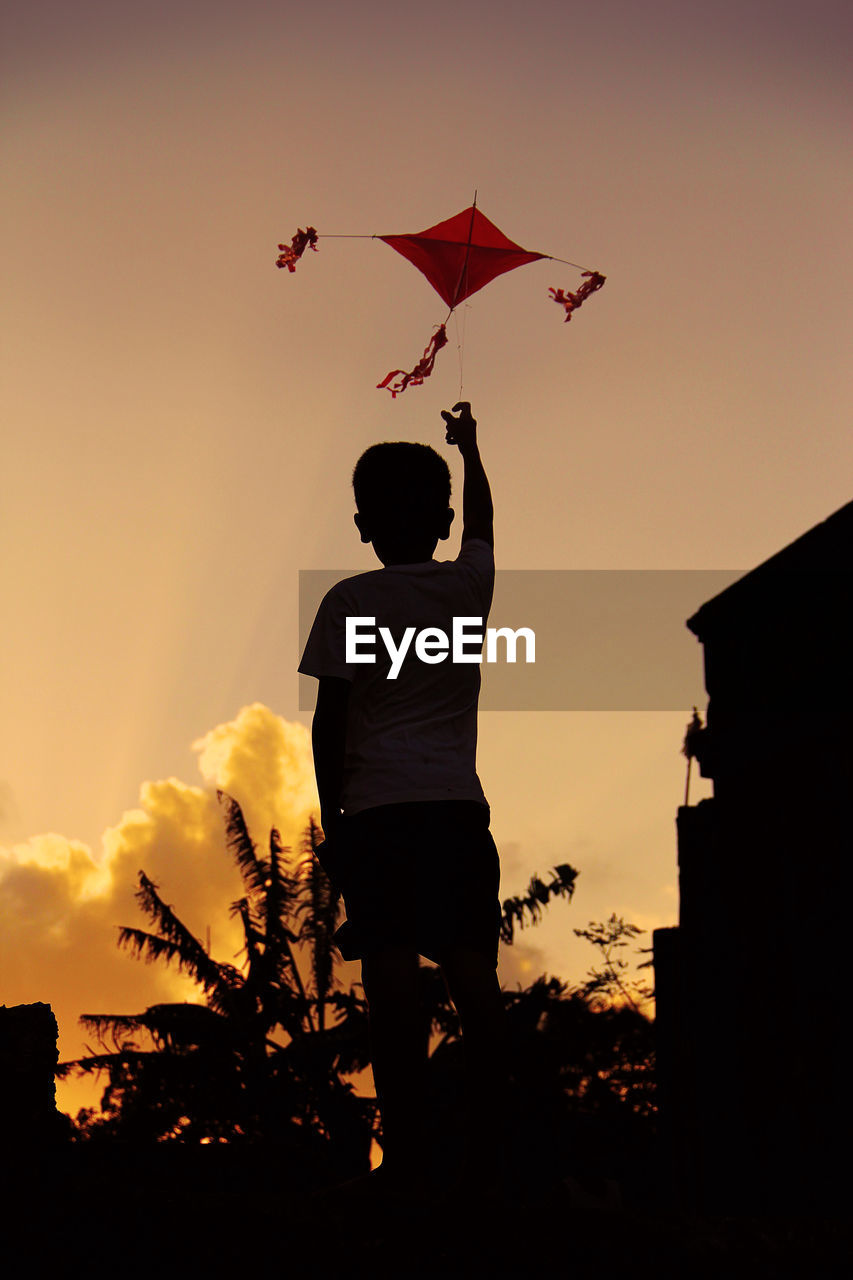 Rear view of silhouette boy flying kite against sky during sunset