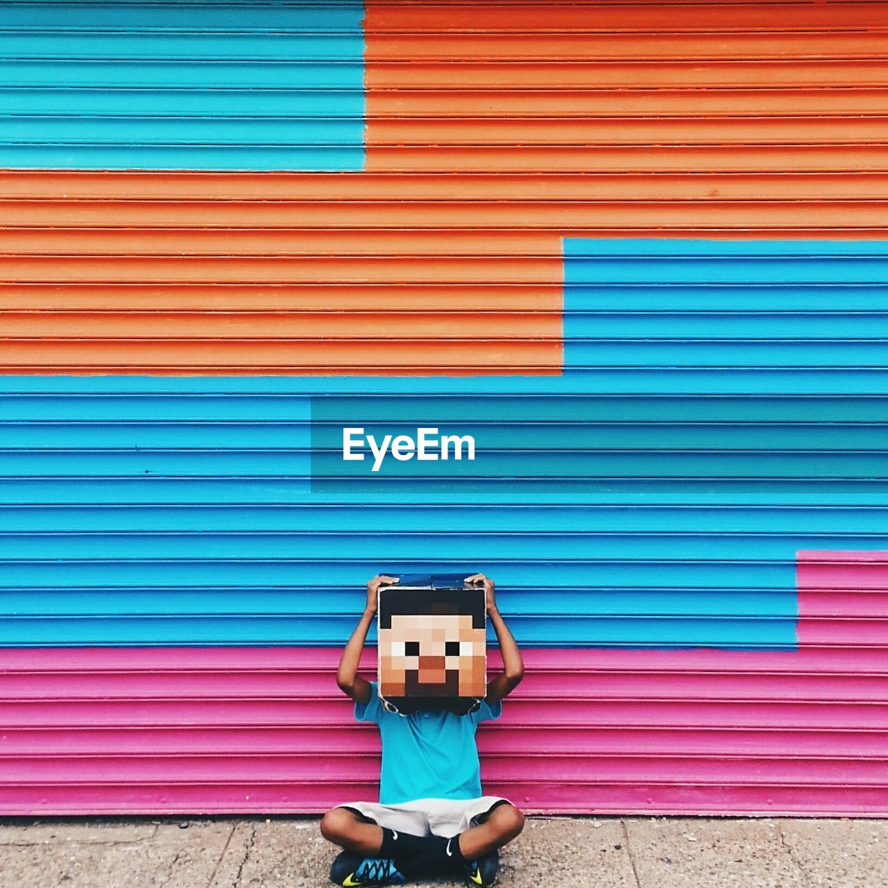 Obscured person sitting with cross-legged and mask against colorful store shutter