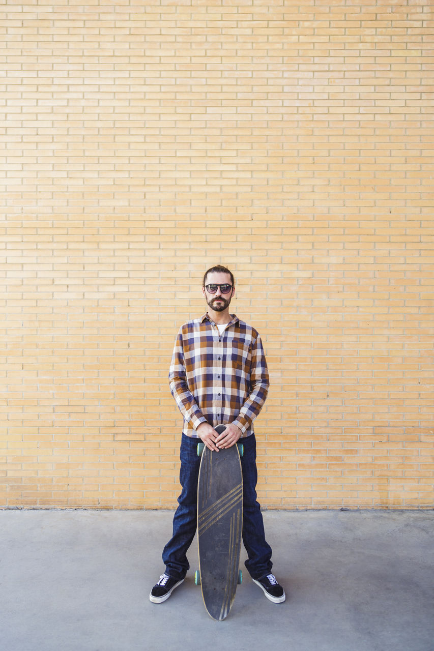 Portrait of man holding skateboard standing against brick wall