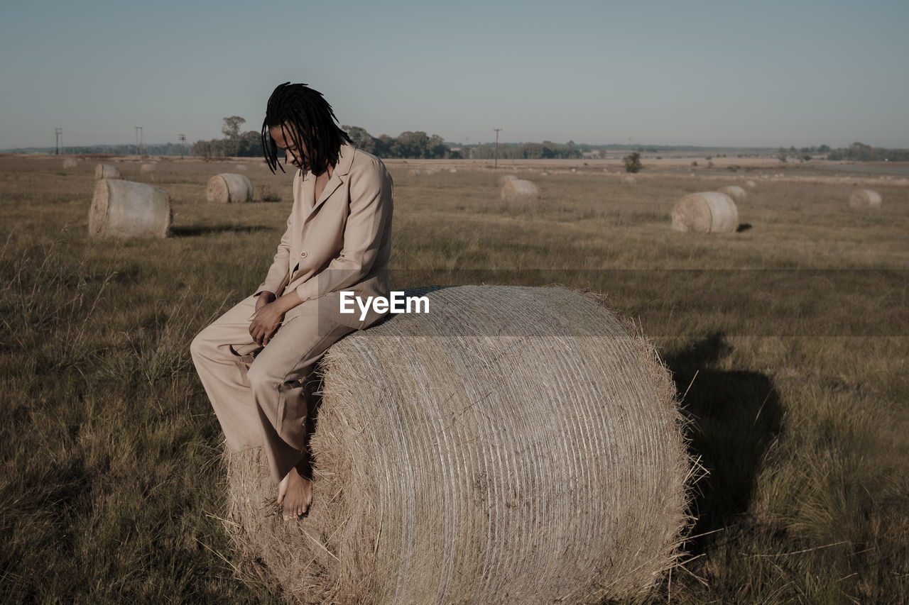 Full length of young man sitting on hay bale at agricultural landscape