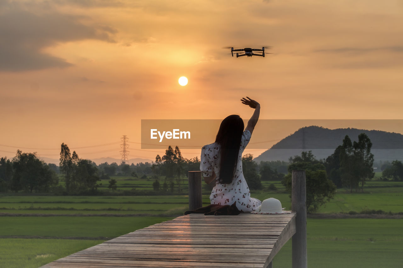 Rear view of woman sitting on footbridge by drone against sky during sunset