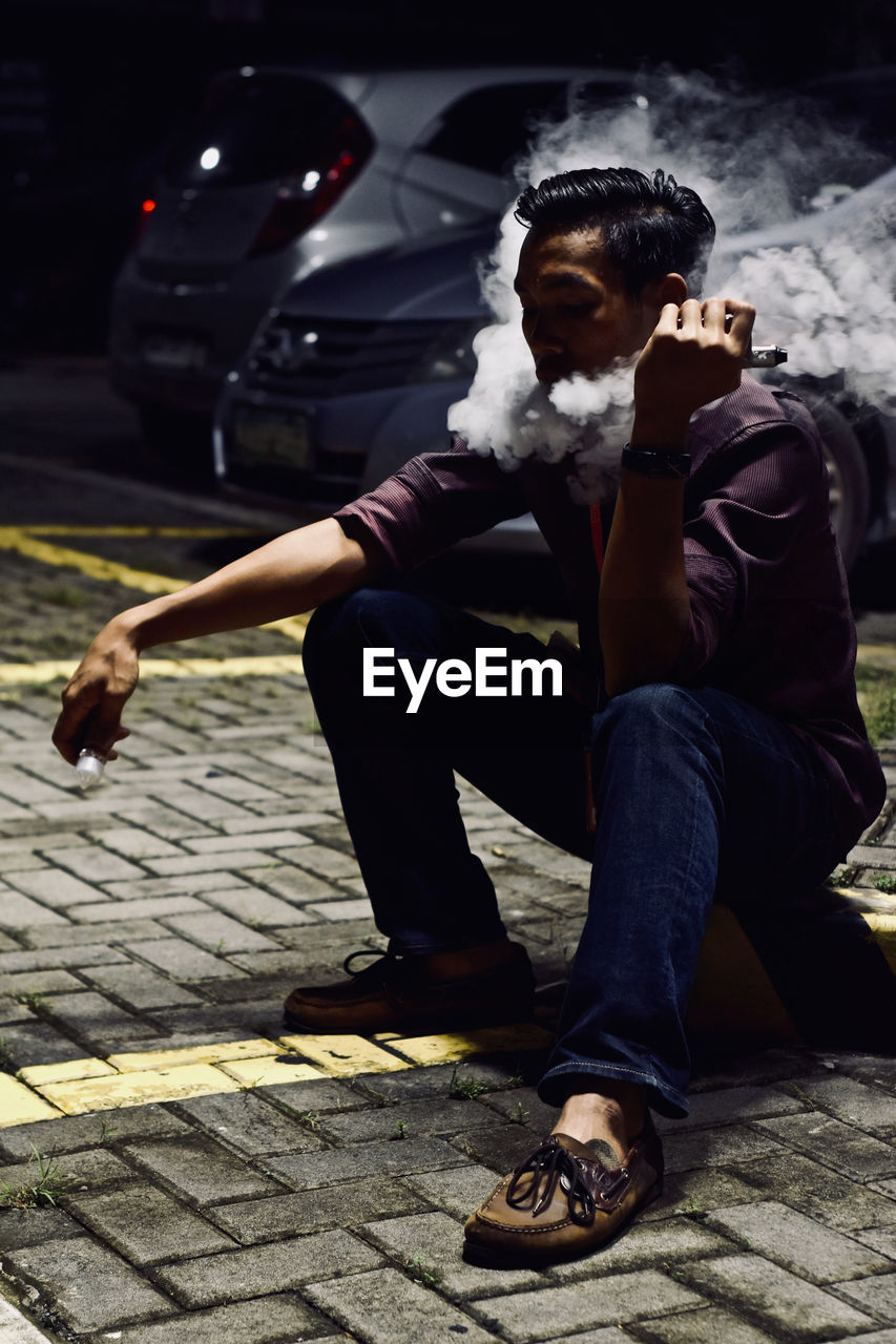 Man exhaling smoke while sitting on sidewalk at night