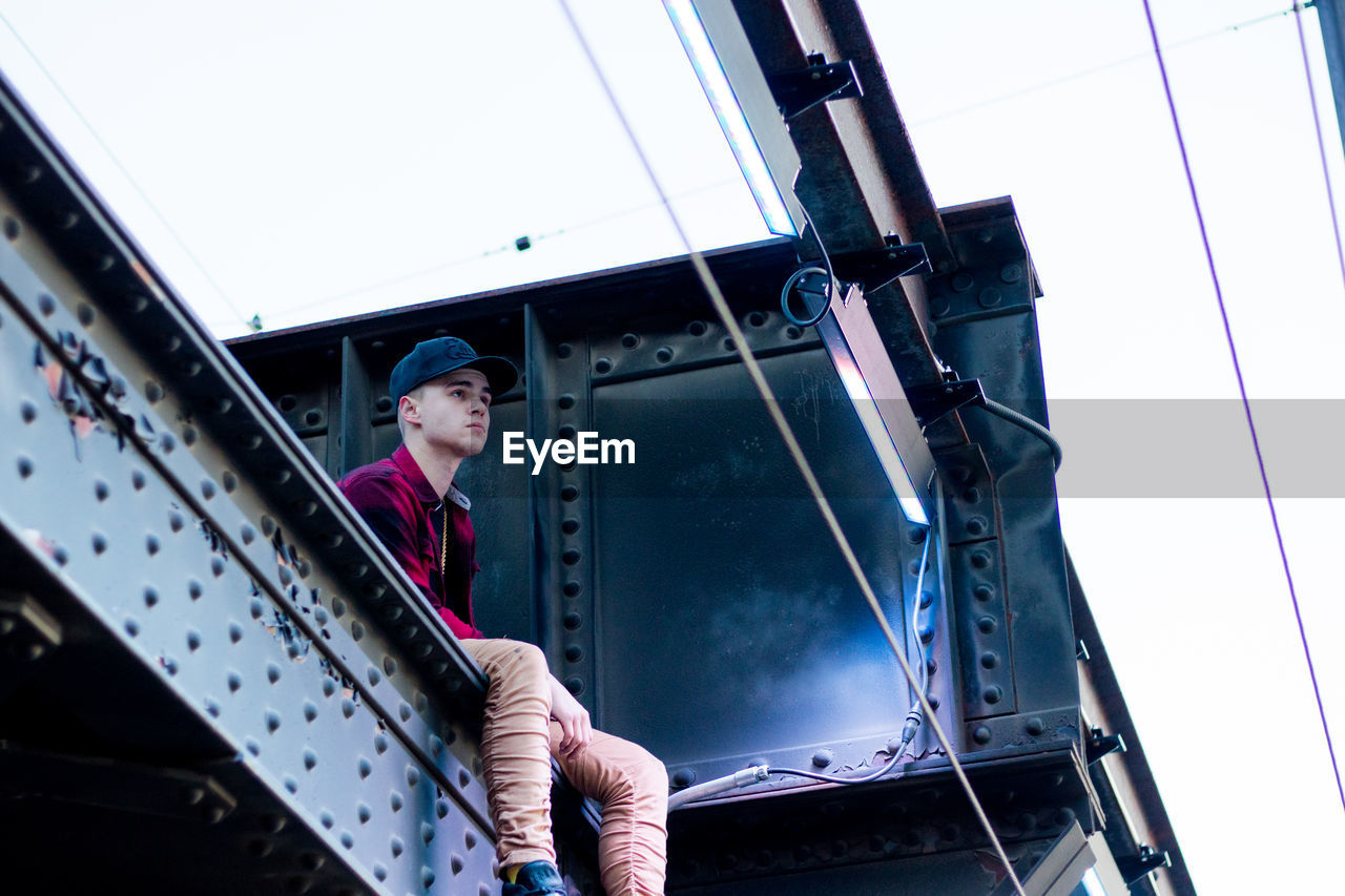 Low angle view of teenager boy sitting on railway bridge against clear sky