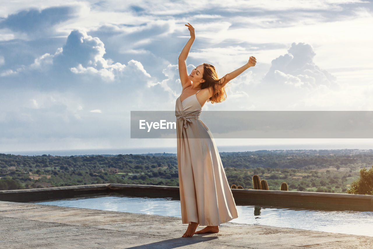 Young brunette woman dancing next to a pool, wearing a cream white dress with a moody sky
