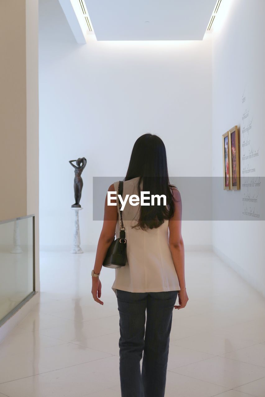 one person, indoors, adult, women, standing, rear view, young adult, long hair, art gallery, hairstyle, full length, lifestyles, clothing, casual clothing, three quarter length, trousers, fashion, person, looking, copy space, exhibition