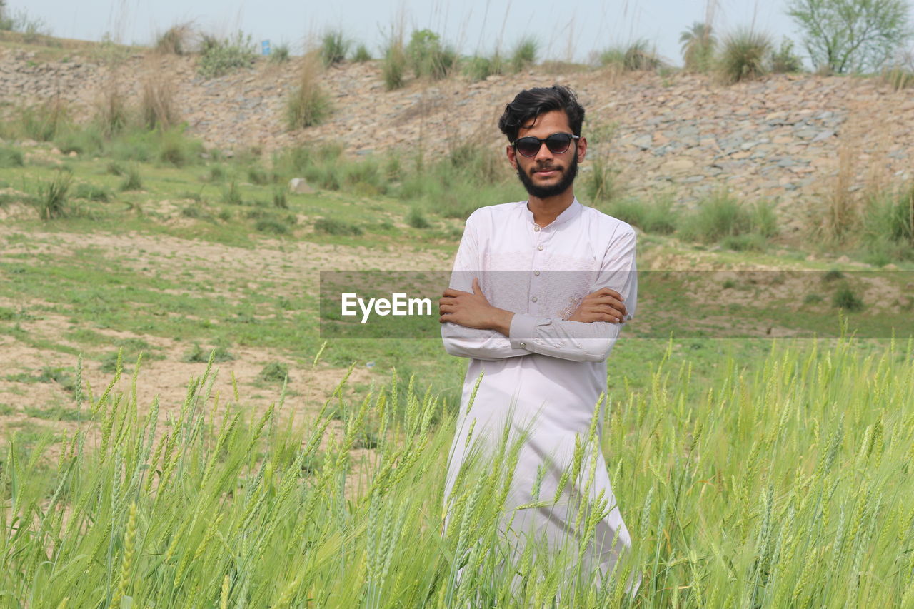 one person, plant, adult, grass, field, nature, land, natural environment, glasses, grassland, fashion, sunglasses, smiling, young adult, men, front view, landscape, agriculture, meadow, portrait, prairie, casual clothing, three quarter length, environment, looking at camera, rural area, day, growth, leisure activity, standing, rural scene, happiness, lifestyles, outdoors, occupation, green, beauty in nature, arms crossed, crop, emotion, facial hair, beard, clothing, tranquility, sunlight, person