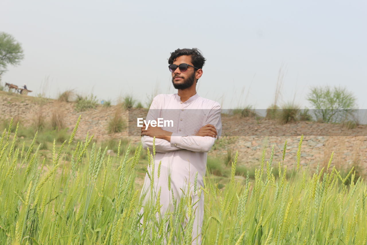 one person, adult, plant, nature, grass, land, men, field, landscape, smiling, natural environment, young adult, agriculture, prairie, rural scene, casual clothing, glasses, sky, portrait, three quarter length, front view, growth, environment, beard, grassland, day, facial hair, meadow, outdoors, fashion, occupation, happiness, crop, leisure activity, standing, rural area, looking, copy space, emotion, looking at camera, sunglasses, lifestyles, relaxation, tranquility, arms crossed, clothing, holding, beauty in nature, waist up