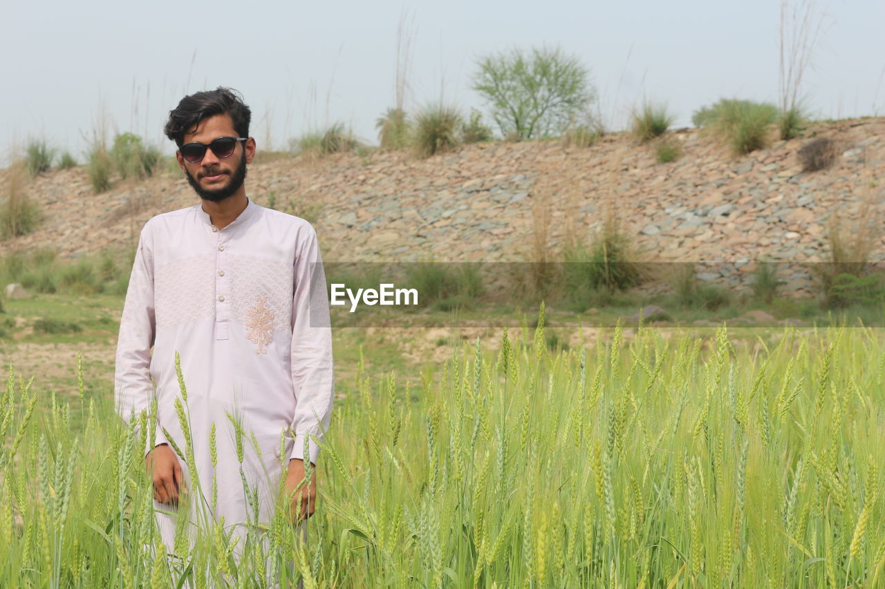 one person, plant, agriculture, field, land, nature, grass, sunglasses, adult, glasses, fashion, prairie, front view, young adult, rural area, grassland, landscape, standing, natural environment, casual clothing, day, crop, portrait, three quarter length, men, rural scene, leisure activity, growth, meadow, sky, smiling, looking at camera, outdoors, lifestyles, clothing, environment, green, beauty in nature, tranquility, waist up, happiness, cereal plant