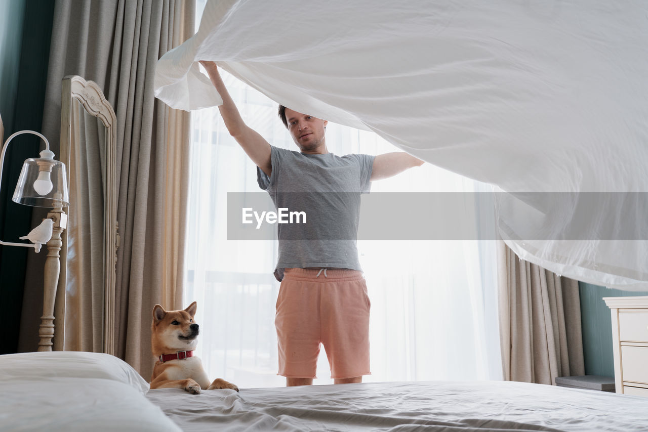 Young man and his funny dog are putting the bedding cover or mattress pad on the bed
