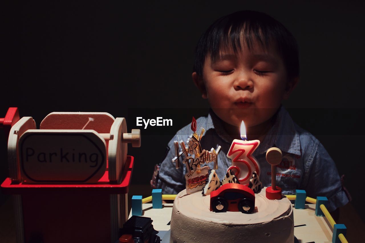Cute boy with eye closed blowing birthday candle on cake in darkroom