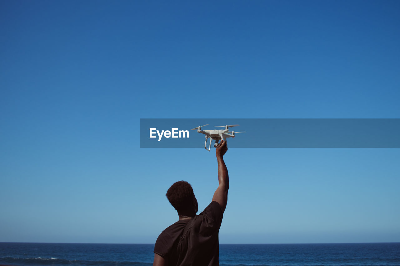 Rear view of man holding drone against blue sky