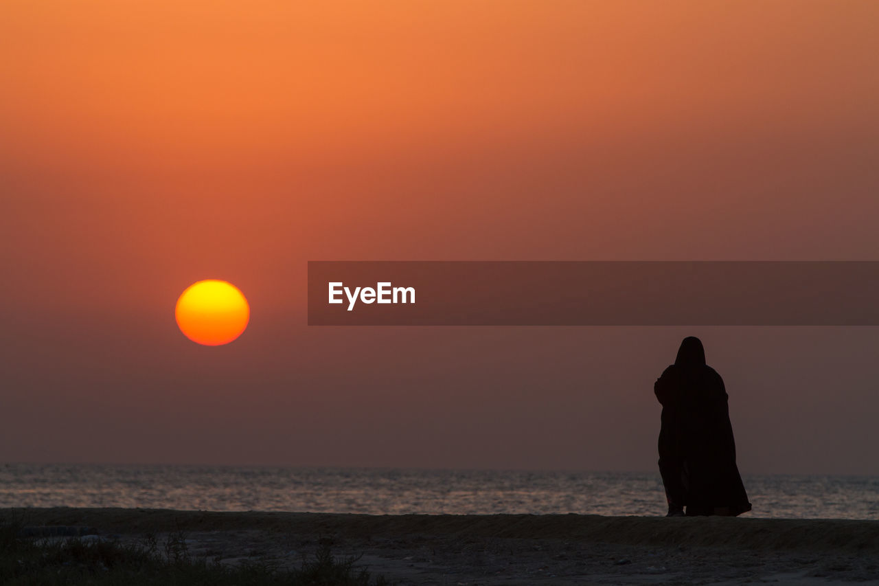 Silhouette woman standing at beach against sky during sunset