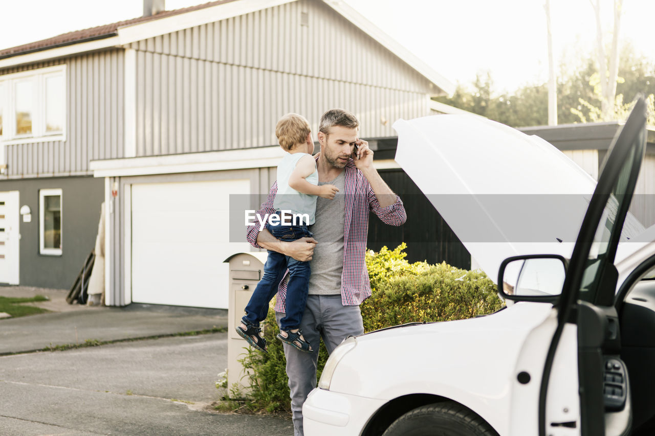 Father carrying son while talking on phone looking at broken down car on street