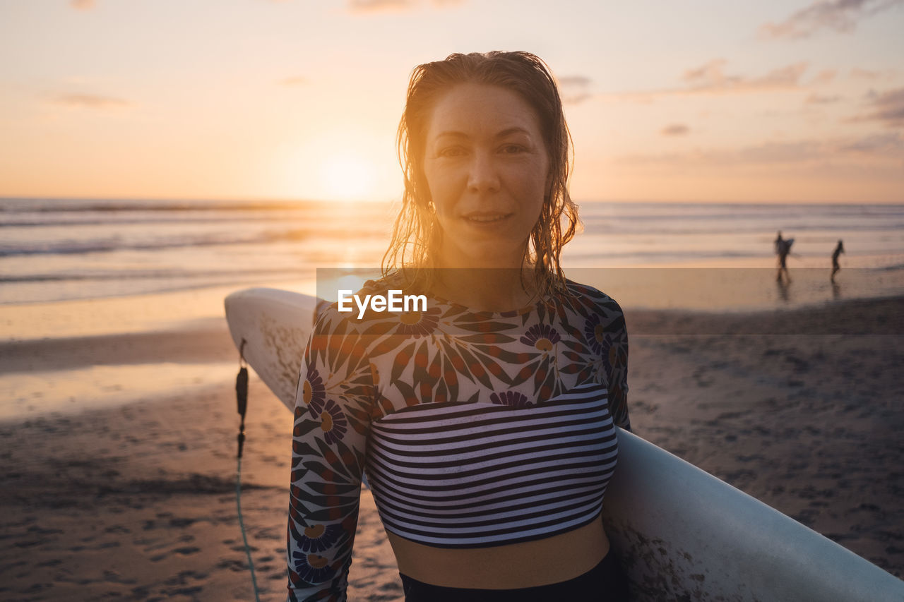 Portrait of mid adult woman with surfboard at beach during sunset