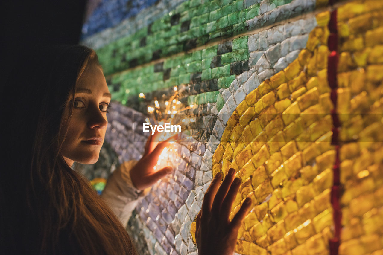 Close-up portrait of young woman with illuminated lighting equipment against wall