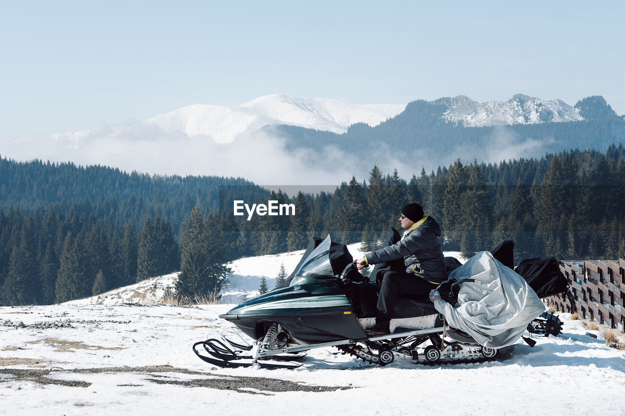 Man sitting on snowmobile in front of snowcapped mountains against sky
