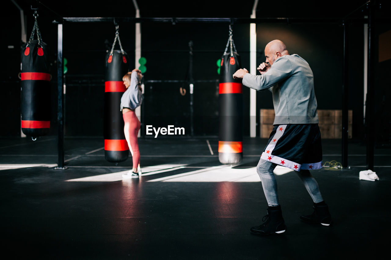 Side view of strong determined sportsman performing boxing exercise during workout in modern sport gym with training woman in background