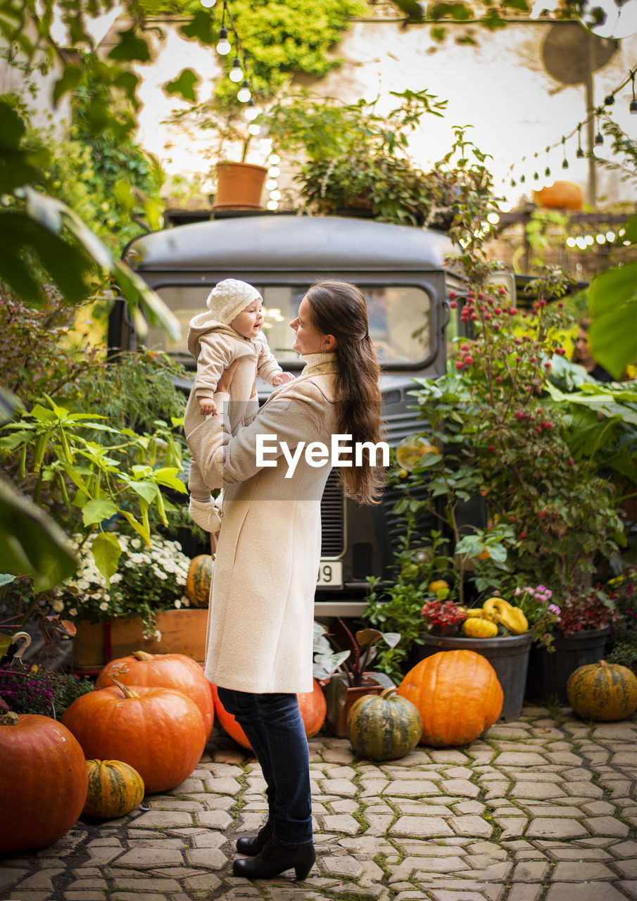 Woman playing with daughter while standing by pumpkins against trees