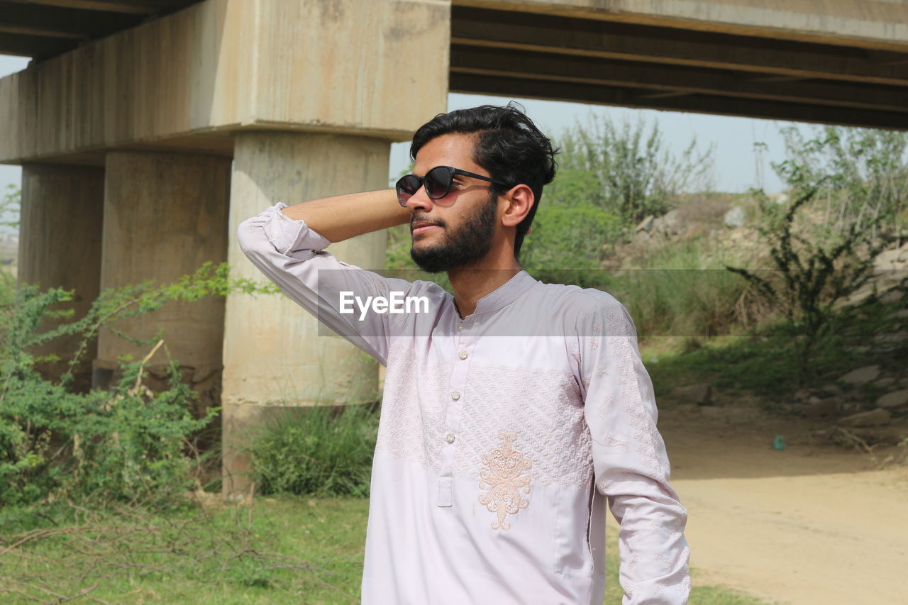 one person, young adult, sunglasses, glasses, adult, standing, fashion, beard, men, casual clothing, facial hair, waist up, nature, lifestyles, leisure activity, day, front view, person, plant, architecture, clothing, outdoors, portrait, built structure, shirt, spring, looking, land, button down shirt, looking away, three quarter length, grass