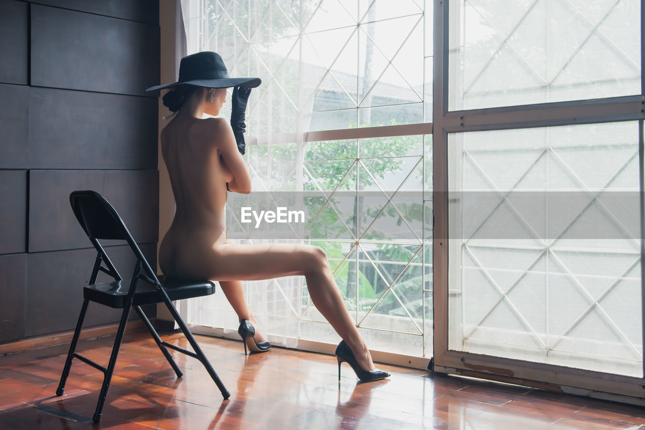 Naked woman sitting on chair by curtain against wall at home