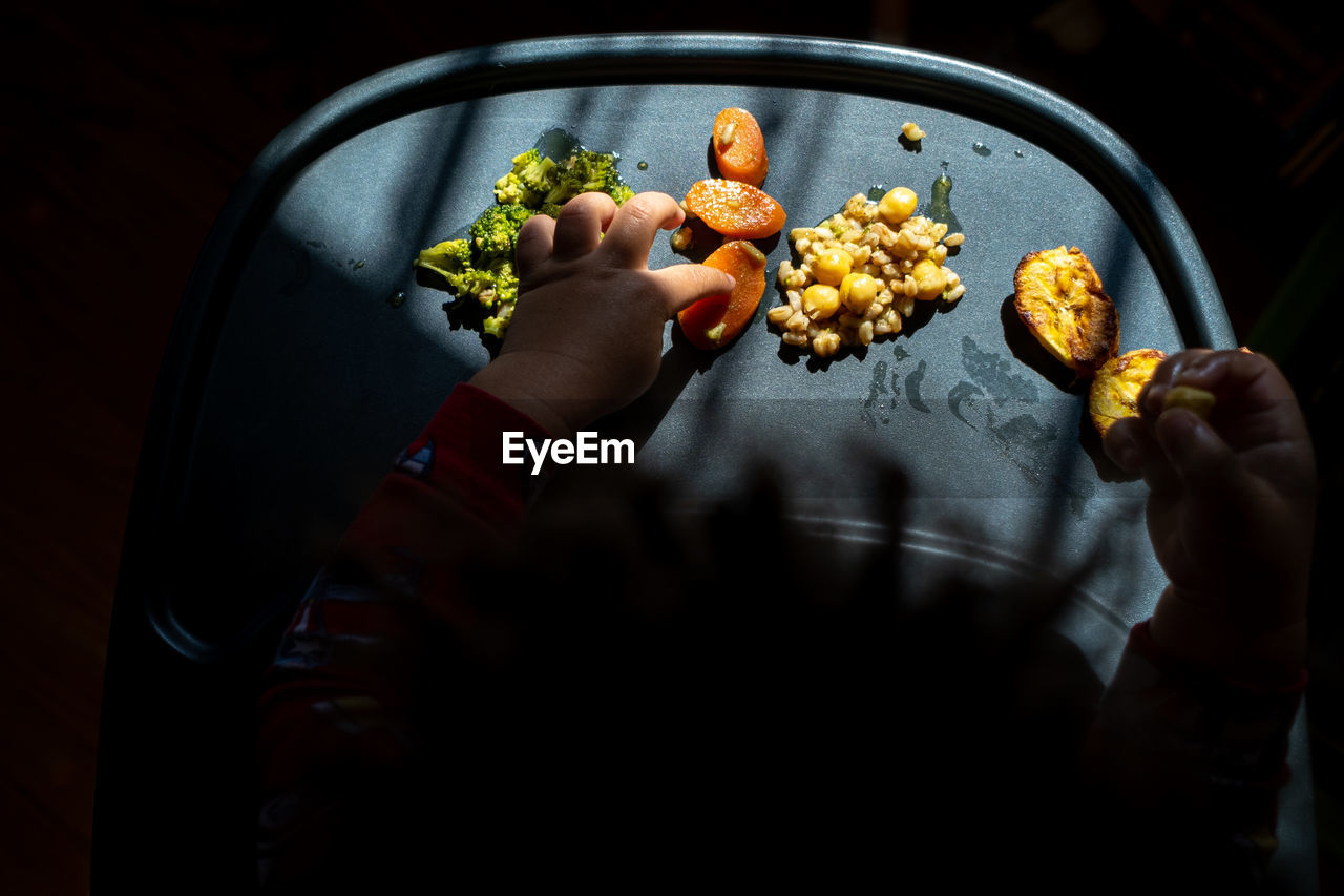 High angle view of a toddler's hands exploring a tray of brightly colored foods during mealtime