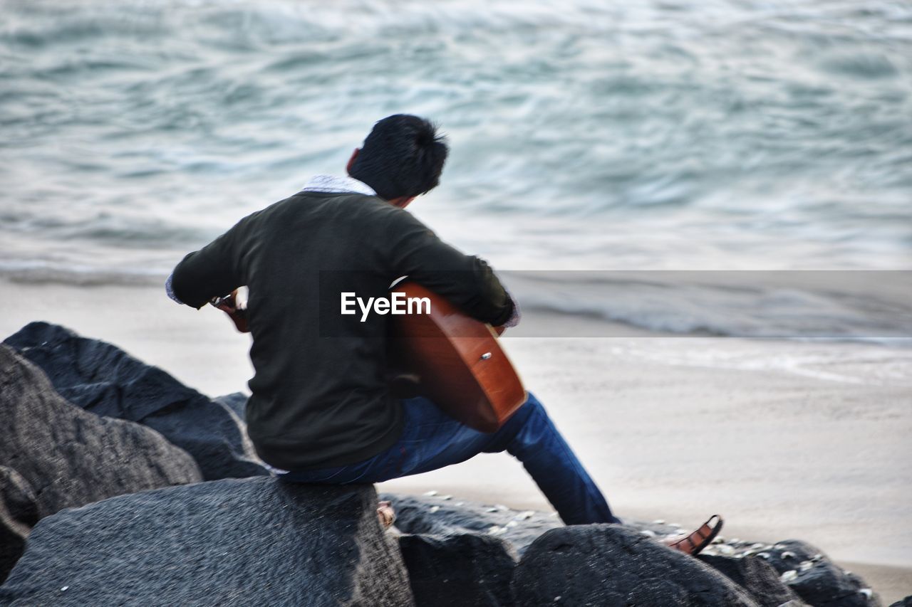 REAR VIEW OF MAN SITTING ON ROCK BY SEA