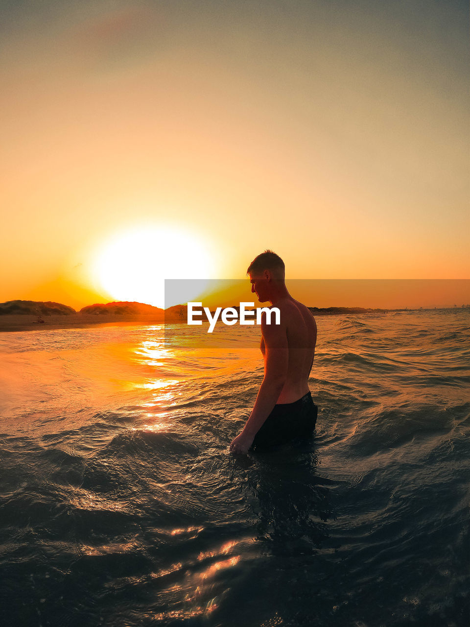Shirtless man standing in sea against sky during sunset