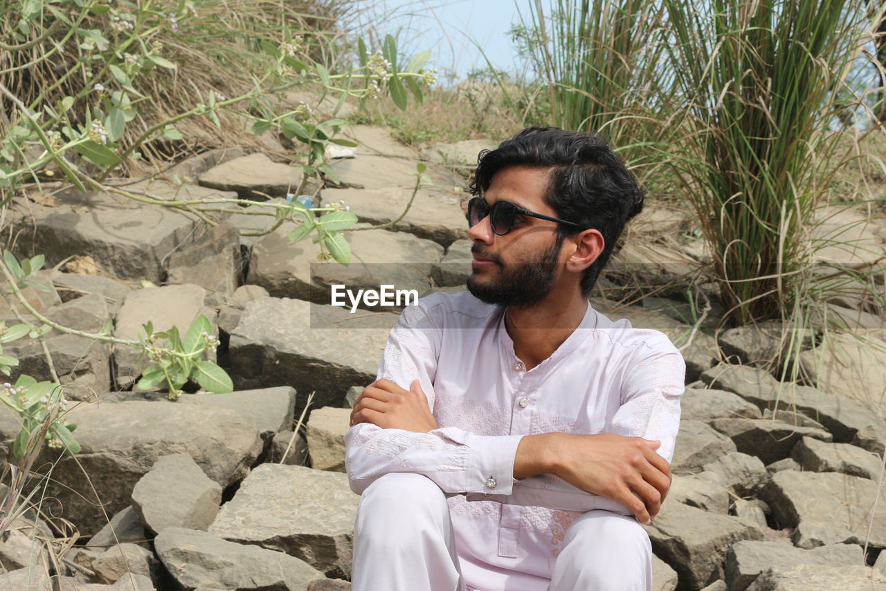 one person, sitting, young adult, nature, men, leisure activity, adult, sunglasses, beard, casual clothing, facial hair, glasses, lifestyles, land, front view, relaxation, day, plant, fashion, rock, person, looking, outdoors, three quarter length, sunlight, looking away, clothing, black hair, vacation, beach, trip, portrait
