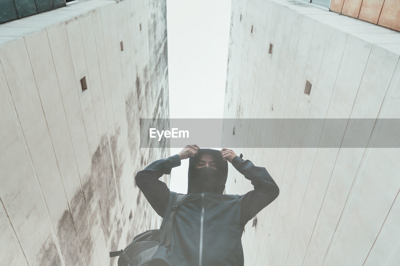 Low angle portrait of man wearing hood standing against buildings