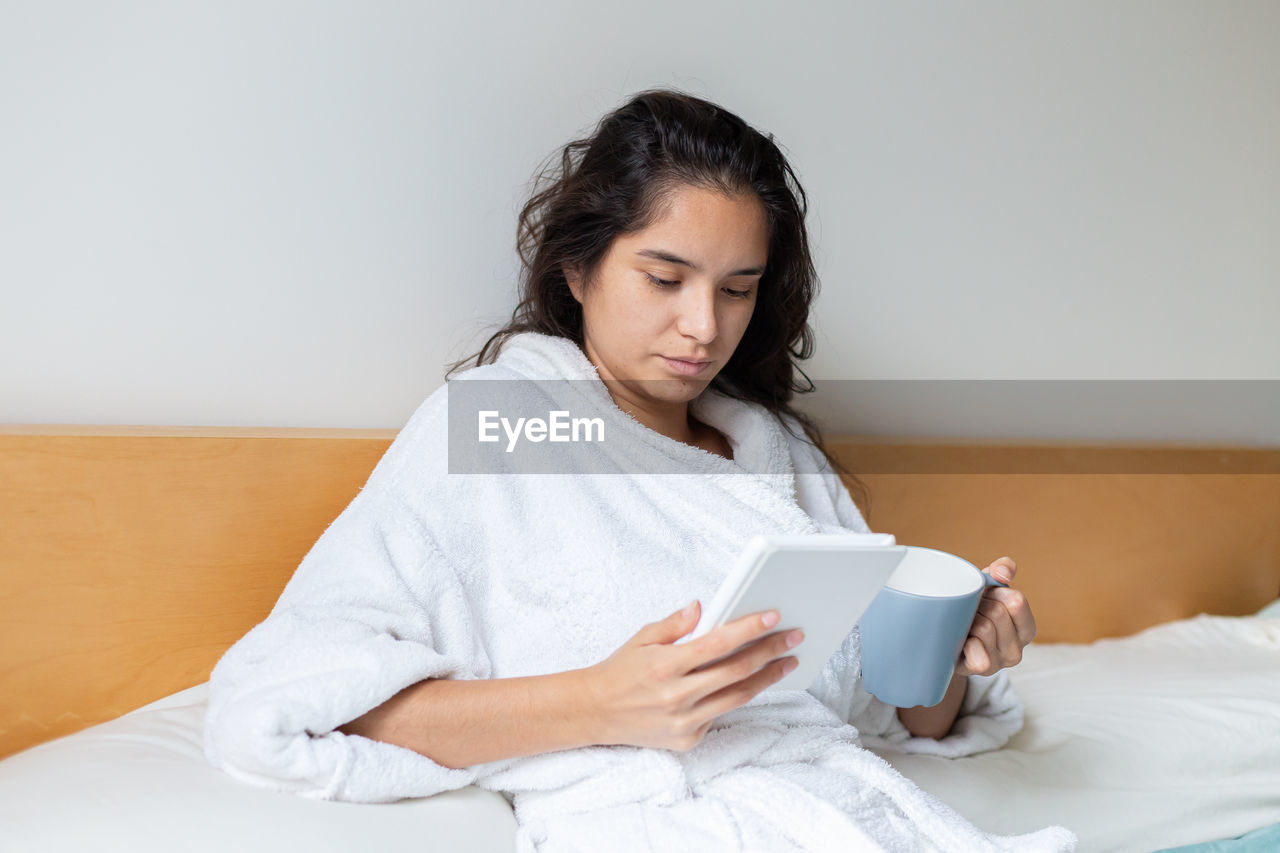 Young woman drinking coffee in bed in bathrobe reading an ebook