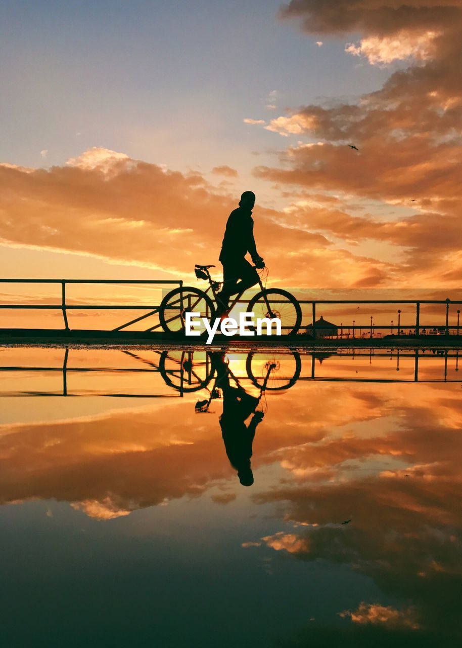 Silhouette man cycling with reflection in lake against sky during sunset