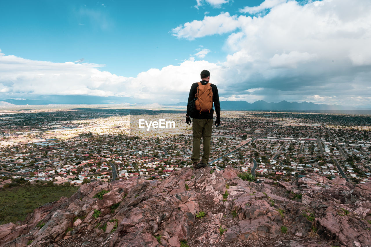 Rear view of male hiker standing on mountain with cityscape in background against cloudy sky