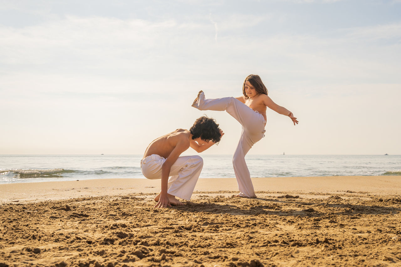 Full length of girl and boy practicing karate at beach against sky
