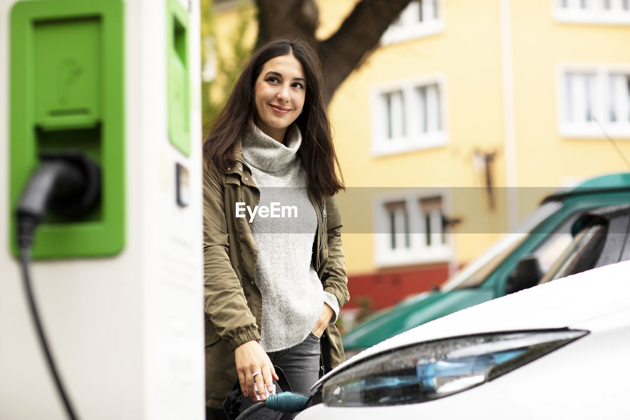 Smiling beautiful woman looking away while standing with hand in pocket and charging electric vehicle at station