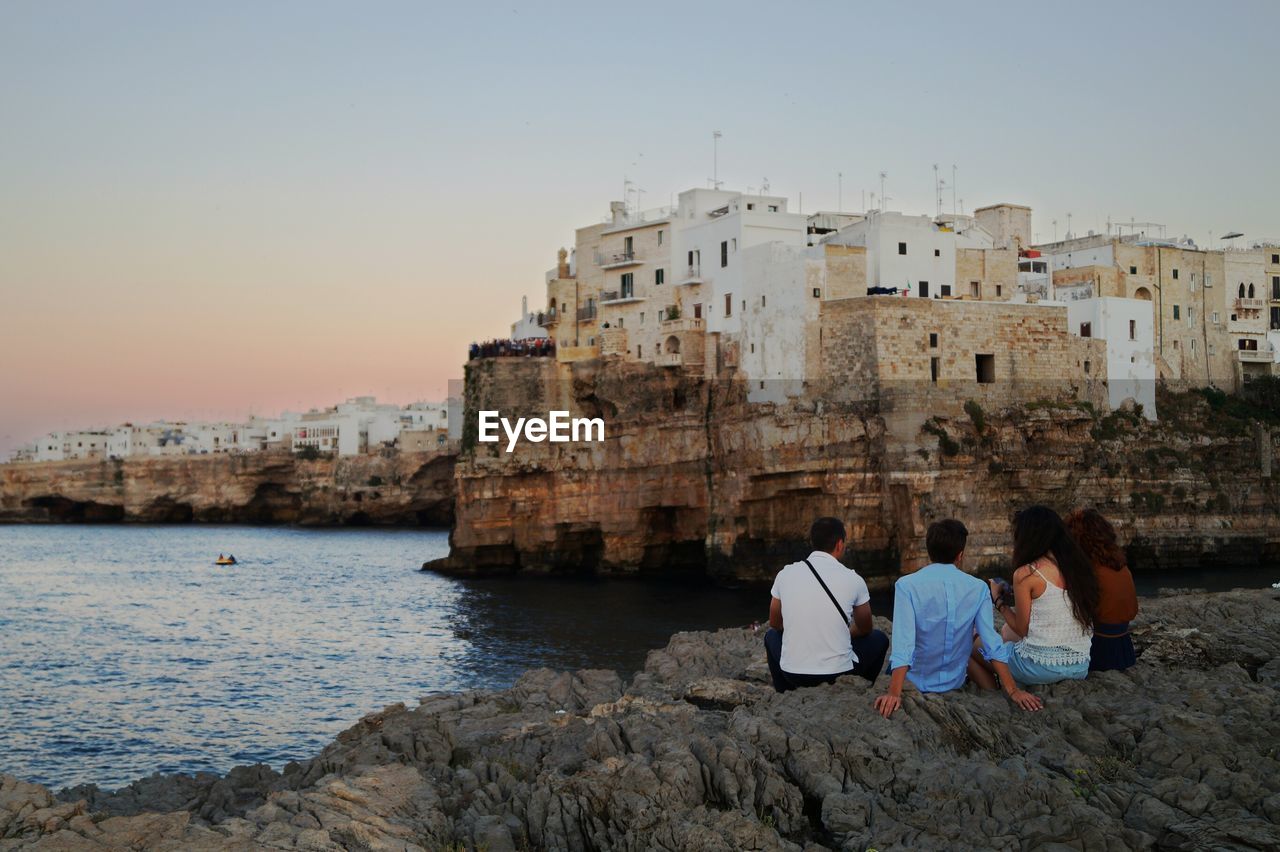 Rear view of friends sitting on rocks at sea shore by buildings against clear sky during sunset