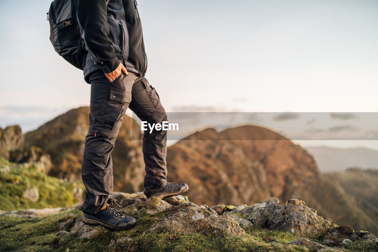 Side view of cropped unrecognizable lonely male explorer with backpack standing on top of green hill and contemplating nature while traveling through mountainous terrain
