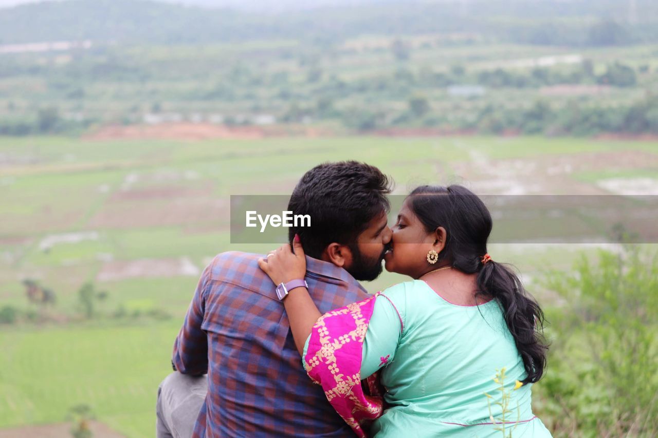love, positive emotion, togetherness, emotion, two people, kissing, adult, embracing, men, women, happiness, affectionate, bonding, landscape, romance, young adult, smiling, ceremony, nature, environment, land, female, rural scene, casual clothing, outdoors, day, portrait, field, person, plant, lifestyles, enjoyment, cheerful, beauty in nature, black hair, grass, waist up, copy space, leisure activity, vacation, arm around, standing