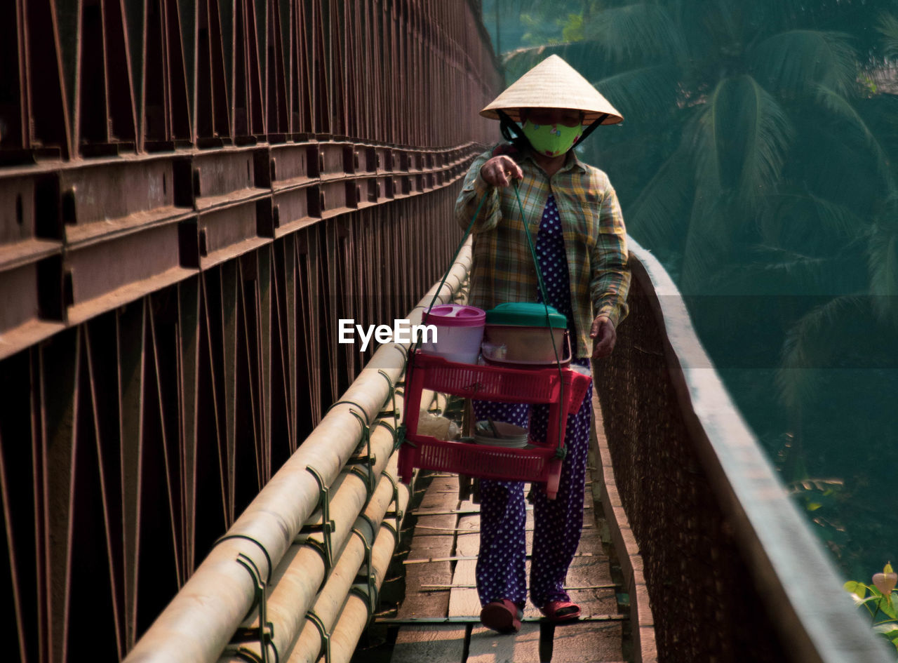 Woman wearing hat carrying containers while walking on footbridge