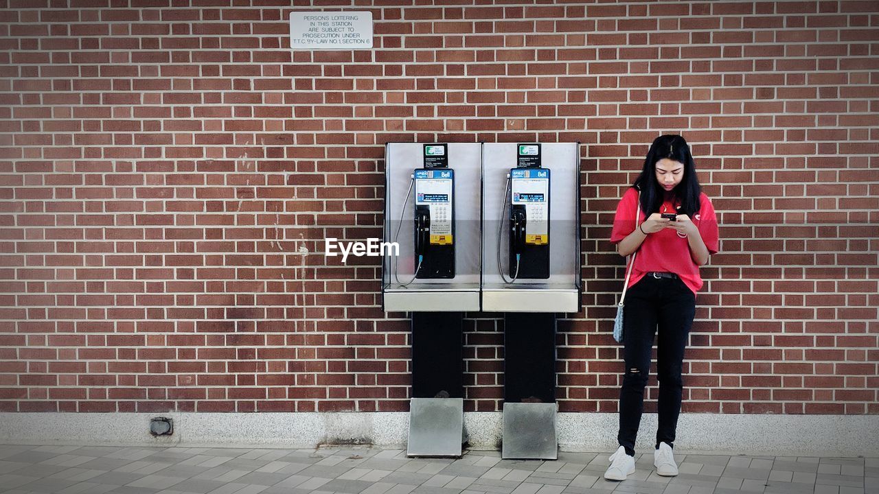 Full length of young woman using phone by telephone booth against brick wall