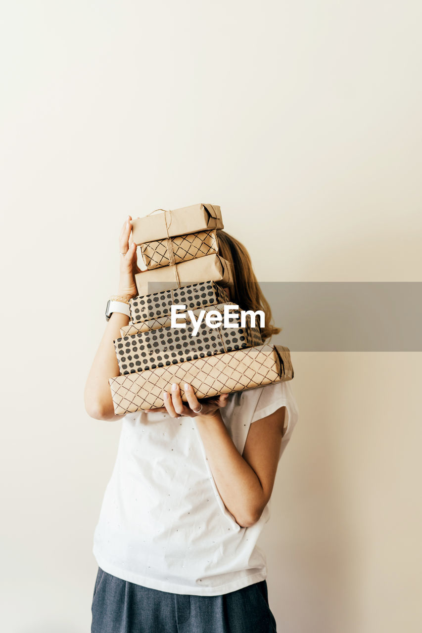 Young cheerful female holds a stack of gift boxes packed in craft paper and hides behind them.