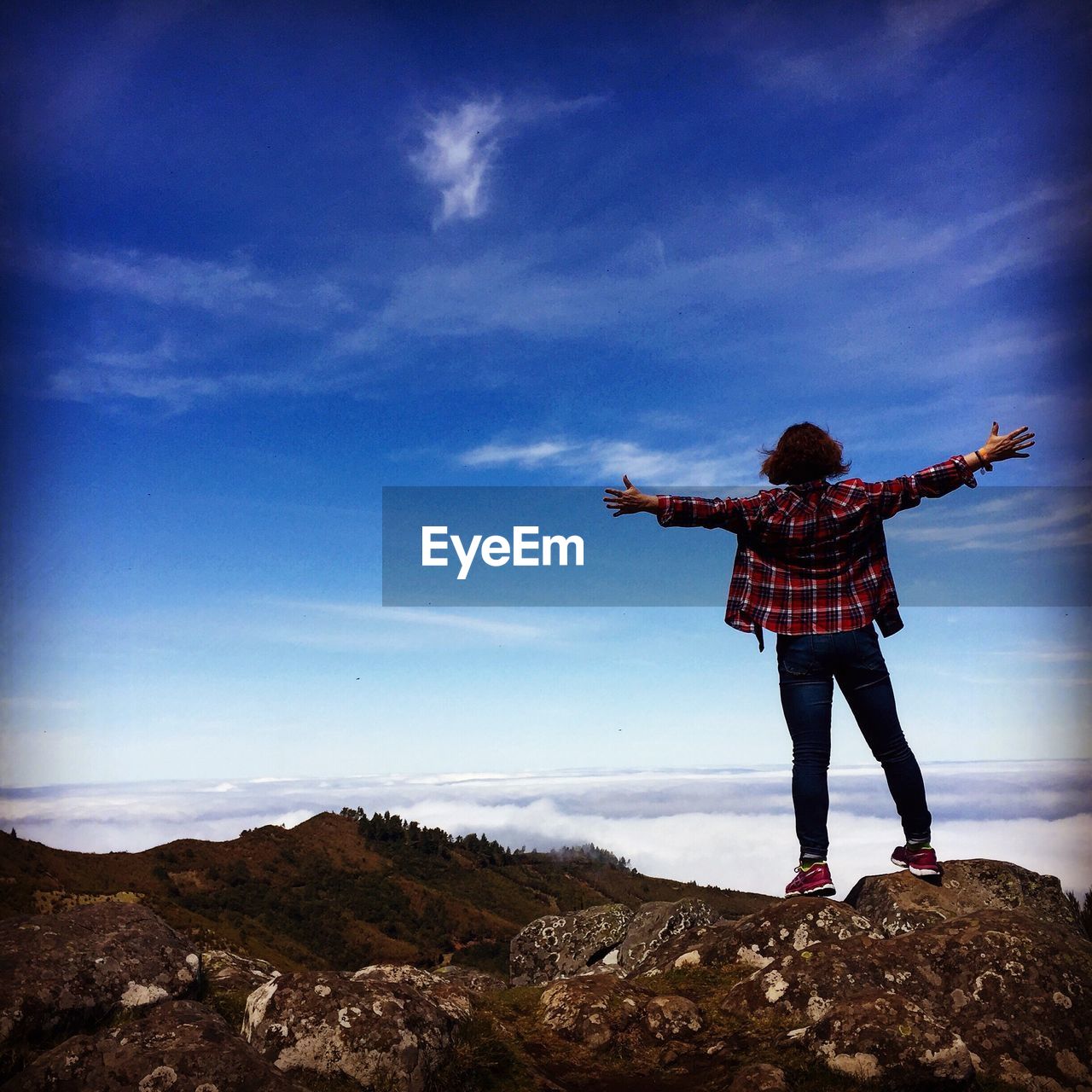 Rear view of woman standing with arms outstretched looking at blue sky from mountain peak