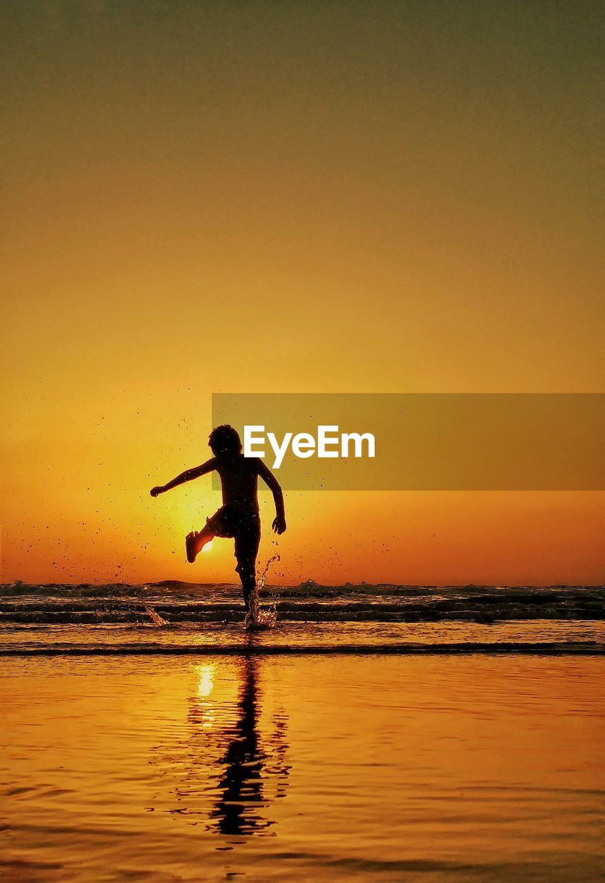 Rear view of silhouette boy enjoying on sea shore against sky during sunset